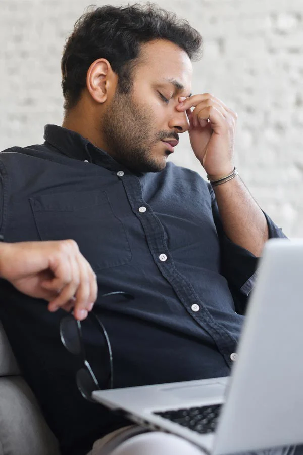 A middle-aged man with a laptop closing his eyes and rubbing his nose. He needs treatment for Adrenal Fatigue Syndrome from Thrive Family Practice & Med Spa in Statesboro.