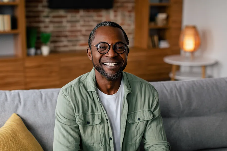 Andropause-Doctor A middle aged black man in a green button up shirt sits on his couch smiling, happy with his treatment for andropause and low testosterone from Thrive Family Practice & Med Spa in Statesboro.