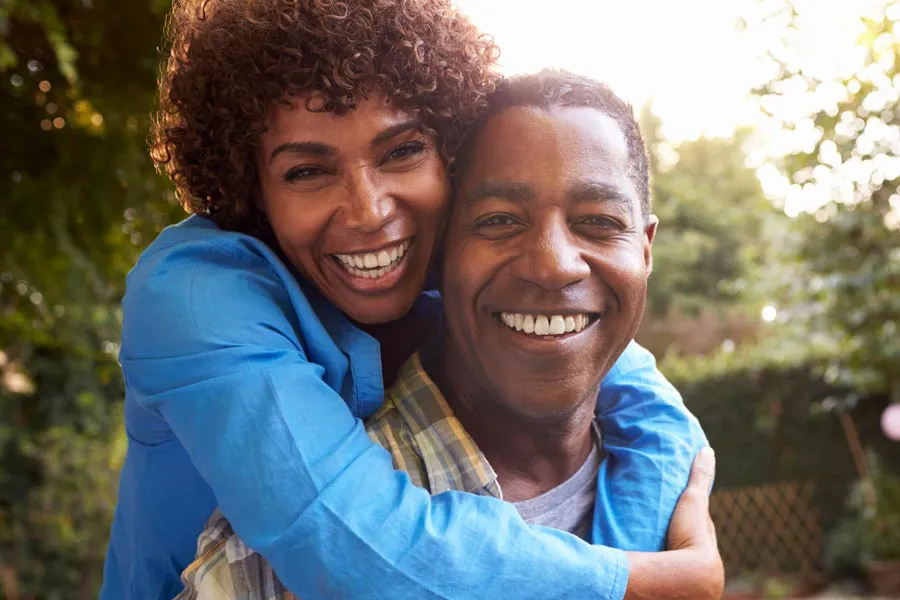 Autoimmune-Doctor A middle-aged African American couple outside, the woman embracing the man from behind pressing her cheek to his, both are smiling due to successful Autoimmune Disorder treatment from Thrive Family Practice & Med Spa in Statesboro.