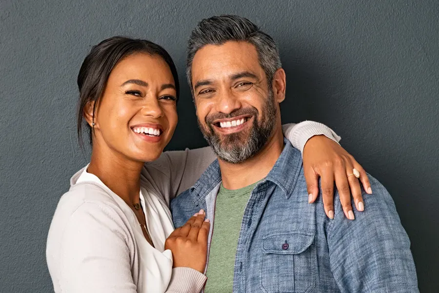 A middle-aged couple with their arms around each other standing against a gray background and smiling, happy with the bioidentical hormone therapy they received from Thrive Family Practice & Med Spa in Statesboro.