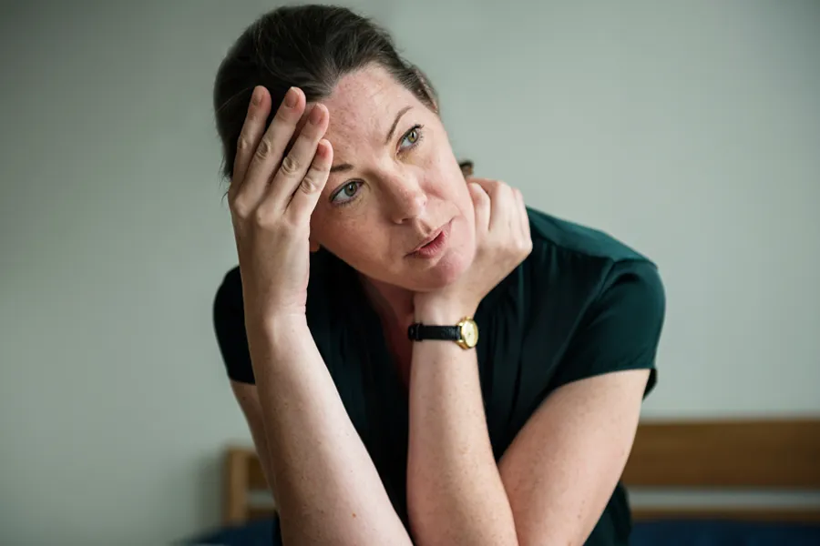 A woman in a dark blue dress sits on her bed holding her head in her hands with an exhausted expression. Get help for chronic fatigue syndrome from Thrive Family Practice & Med Spa in [AERA].