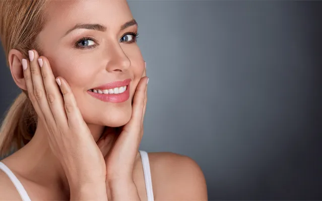 A middle-aged blonde woman with beautiful skin smiles and holds her face. Get facial treatments from Thrive Family Practice & Med Spa in Statesboro.