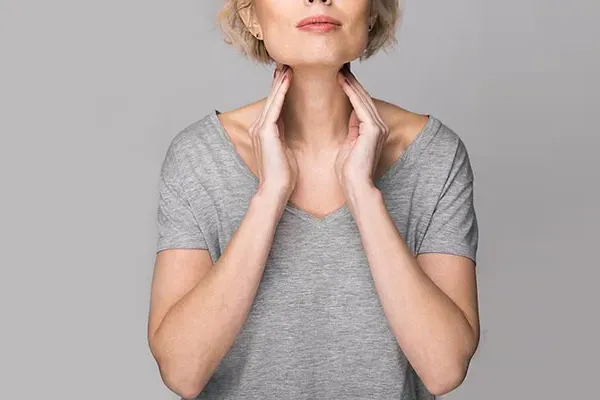 Hashimotos-Thyroiditis-Doctor An older blonde woman in a gray t-shirt holds her hands to her neck checking for signs of thyroid problems. Get help for Hashimoto's Thyroiditis from Thrive Family Practice & Med Spa in Statesboro.