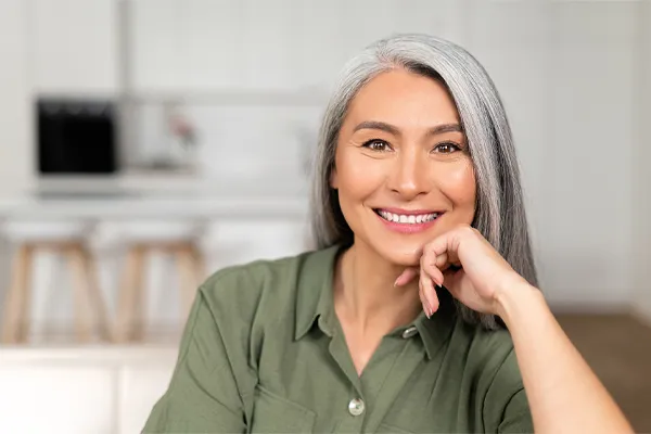 Menopause-Clinic An asian woman with long gray hair and a green shirt sitting and smiling on her couch, happy with her menopause care from [NAME} in Statesboro.