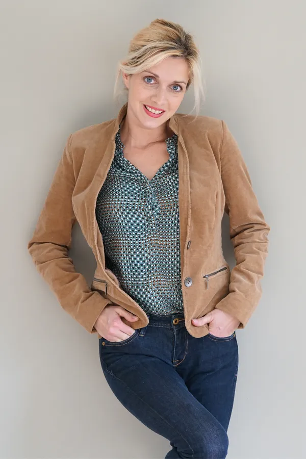 A smiling blonde woman in a tan leather jacket leans against a gray wall. Schedule metabolic syndrome from Thrive Family Practice & Med Spa in Statesboro.