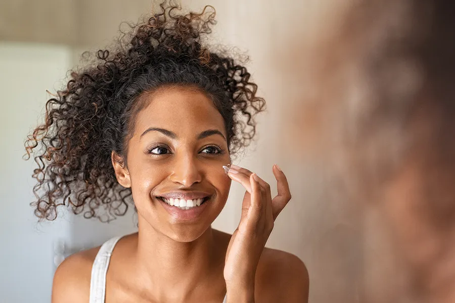 Neova-Smart-Skincare-Clinic A beautiful mixed-race woman looks in the mirror smiling and applies skin cream to her cheek. Get Neova Smart Skincare from Thrive Family Practice & Med Spa in Statesboro.