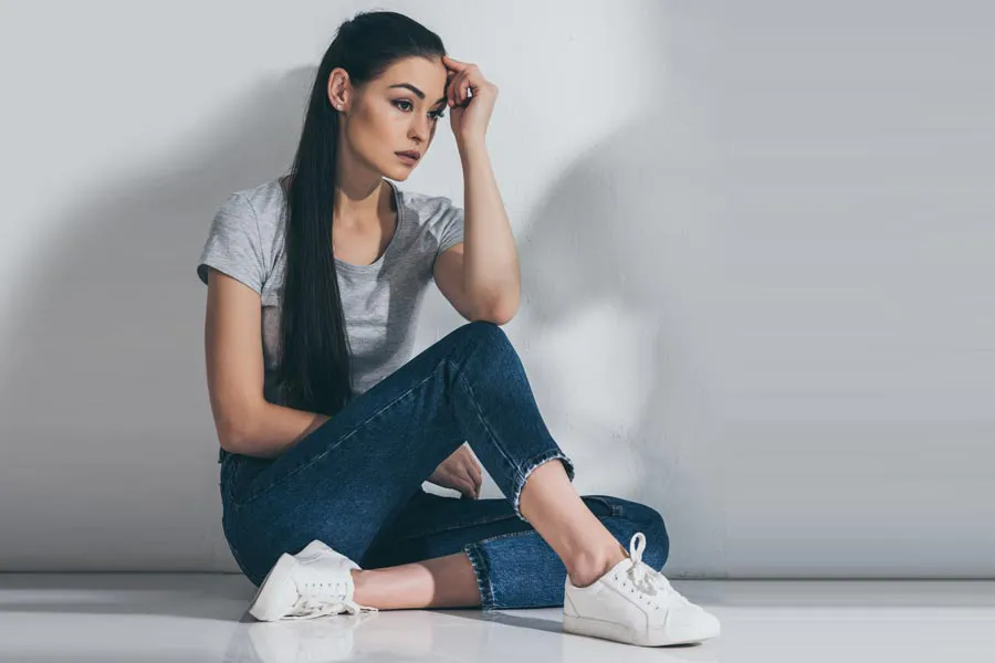 PCOS-Doctor A young brunette woman in a gray t-shirt and jeans sits against a gray wall with her head resting on her hand and a distressed expression. Get help for Polycystic Ovarian Syndrome (PCOS) from Thrive Family Practice & Med Spa in Statesboro.