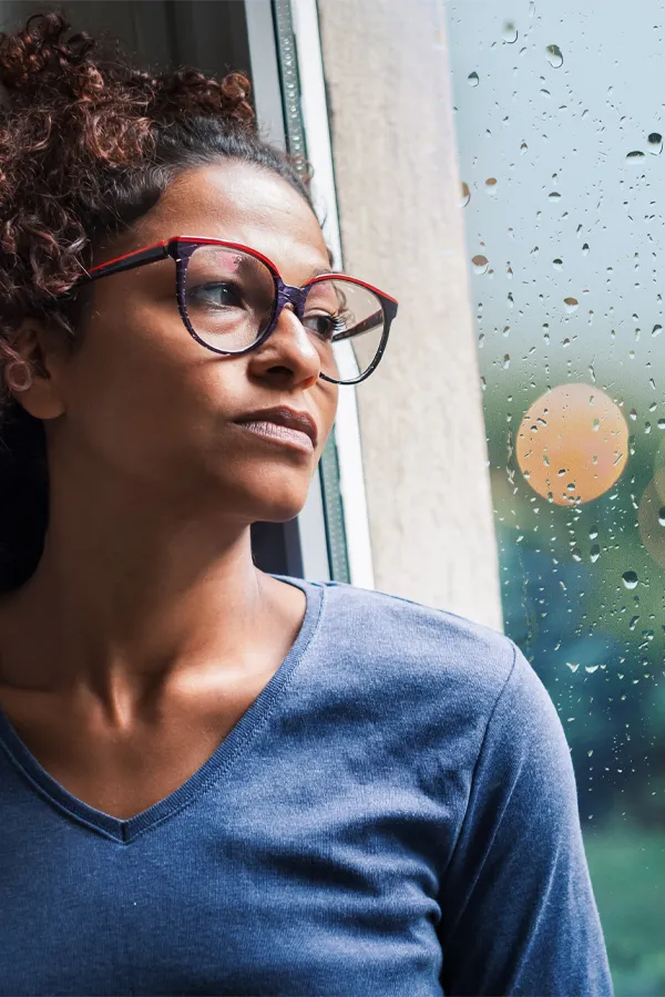 PMDD-Treatment A black woman with with a somber expression and glasses and a gray shirt stares out the window on a rainy day. Get treatment for Premenstrual Dysphoric Disorder from Thrive Family Practice & Med Spa in Statesboro.