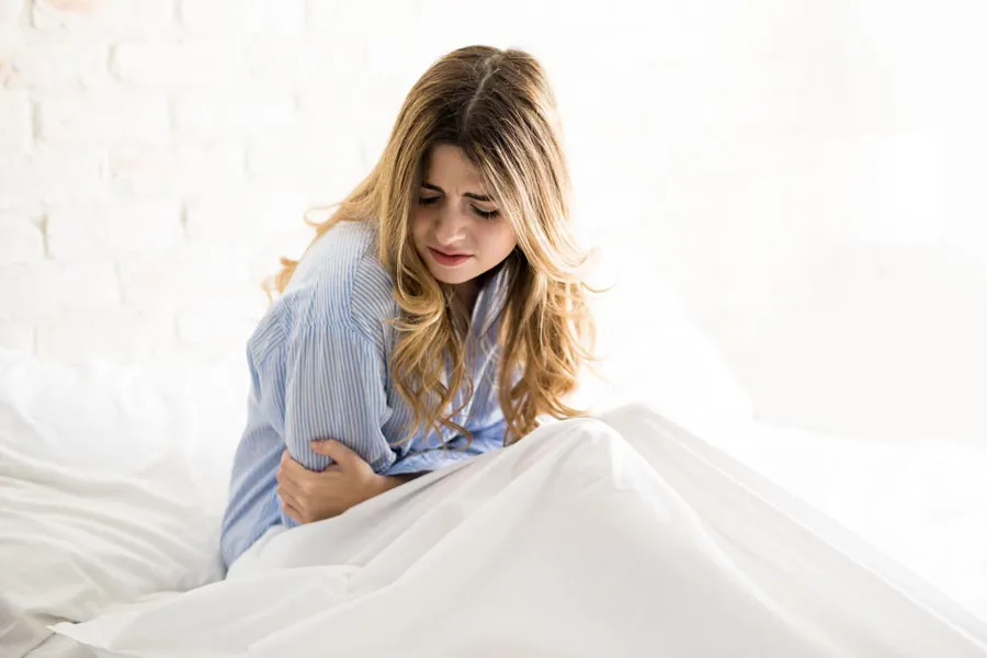 PMS-Therapy A woman with long blonde hair sits in her bed holding her stomach in pain, considering getting care for PMS from Thrive Family Practice & Med Spa in Statesboro.