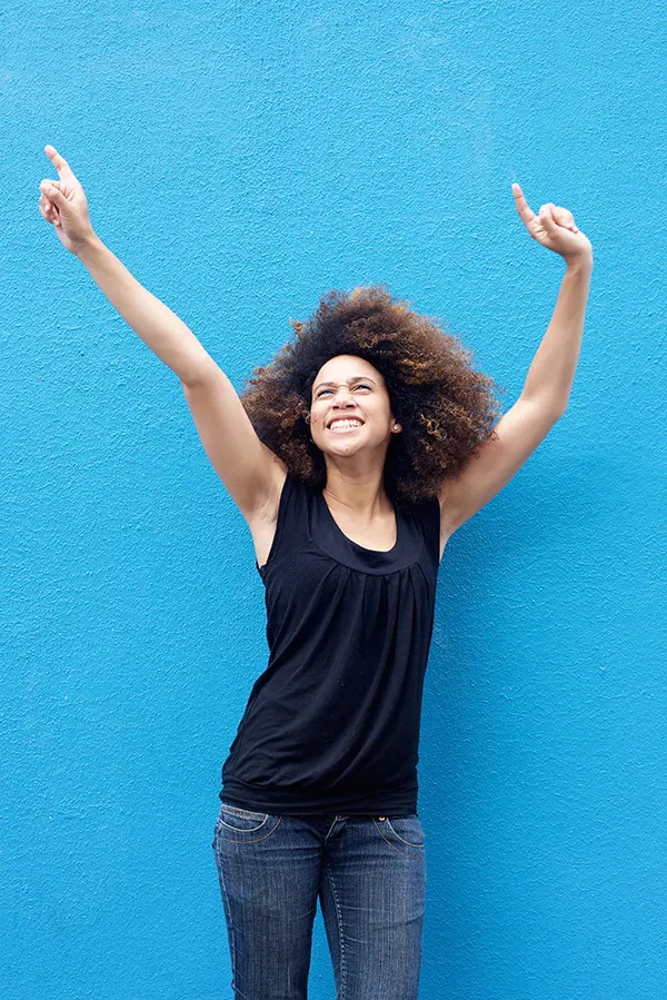 PMS-Treatment A woman in a dark blue tank top standing in front of a bright blue wall, raising her arms in celebration of relief from PMS from Thrive Family Practice & Med Spa in Statesboro.