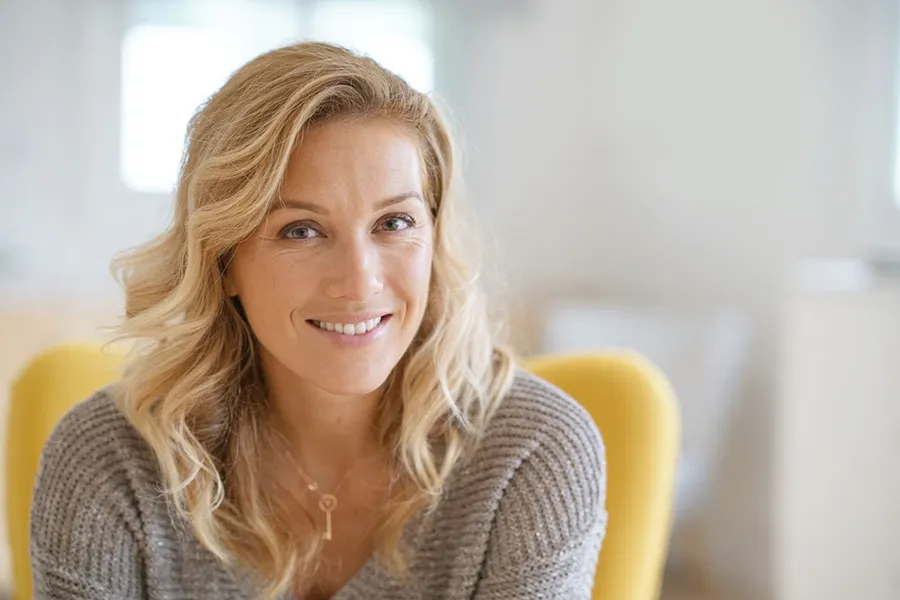 Perimenopause-Doctor A blonde woman wearing a a gray sweater sits inside in a yellow chair, smiling and happy with her perimenopause treatment from Thrive Family Practice & Med Spa in Statesboro.