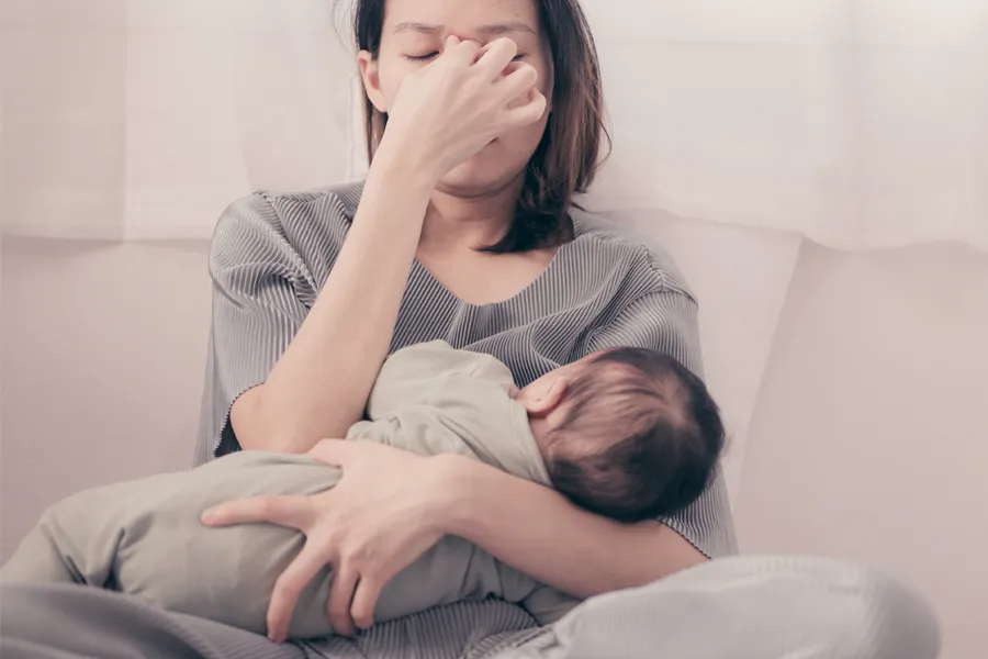 Postpartum-Hormone-Imbalance-Clinic A woman nursing an infant on couch rubs her eyes with exhaustion. Get treatment for postpartum hormone imbalance from Thrive Family Practice & Med Spa in Statesboro.
