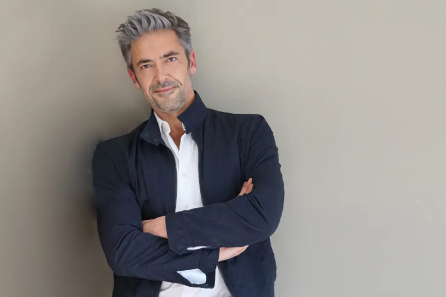 A middle-aged man in a white shirt and dark blue jacket leans against a gray wall smiling. Get testosterone for Low T from Thrive Family Practice & Med Spa in Statesboro.