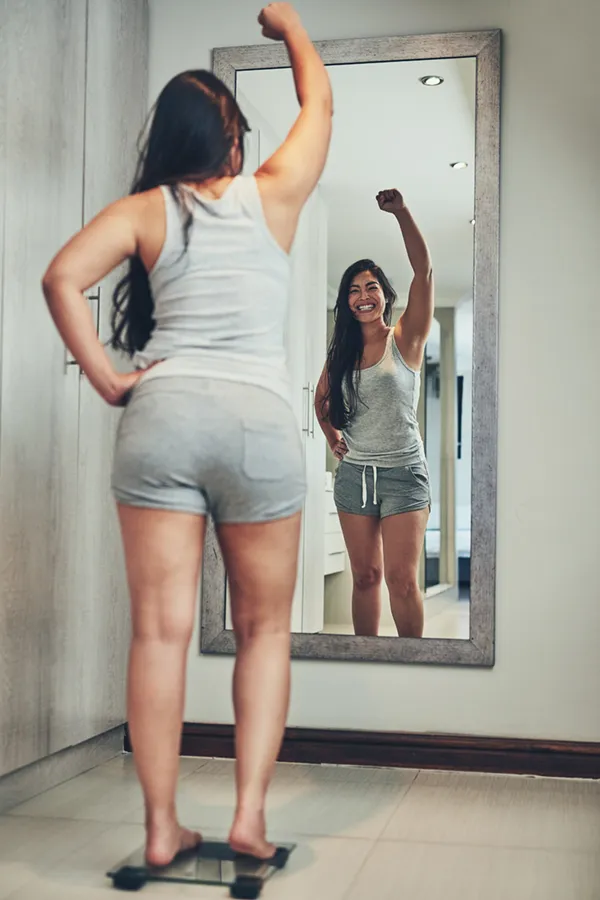 Tirzepatide-Treatment A woman in workout clothes stands on a scale looking in the mirror and pumping her hands in celebration of her successful Tirzepatide treatment from Thrive Family Practice & Med Spa in Statesboro.