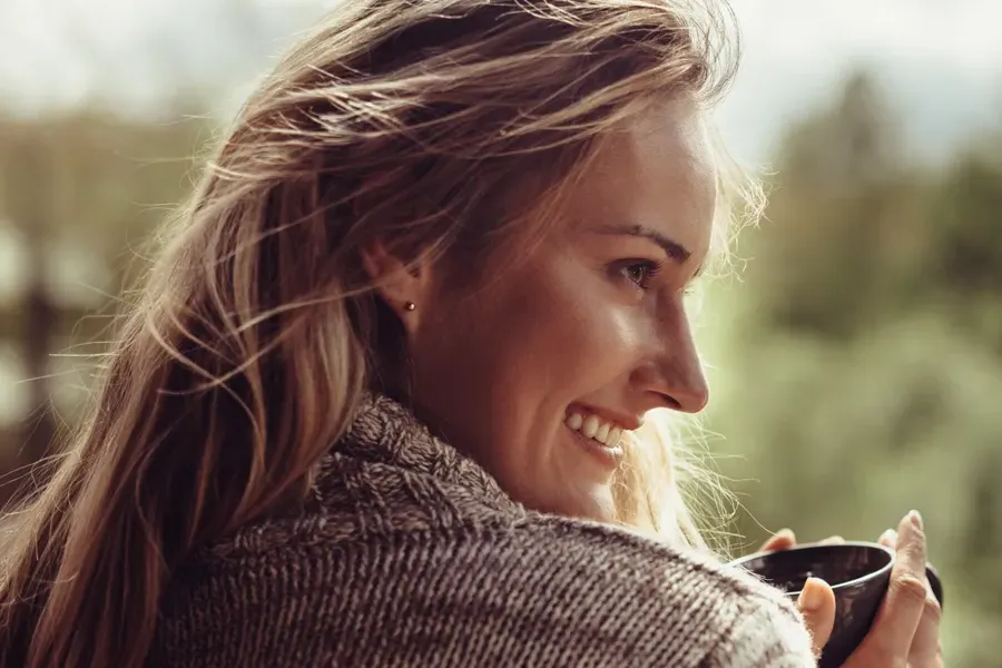 A middle-aged blonde woman in a sweater drinks tea, framed by a forest horizon. Get comprehensive women's health exams from Thrive Family Practice & Med Spa in Statesboro.